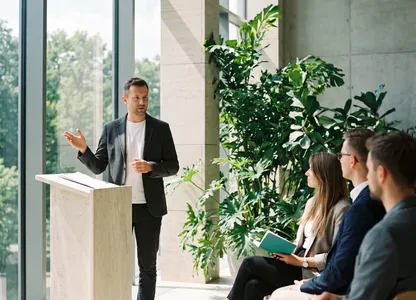 Prelegent przy wysokim oknie mówi do niewielkiej grupy słuchaczy